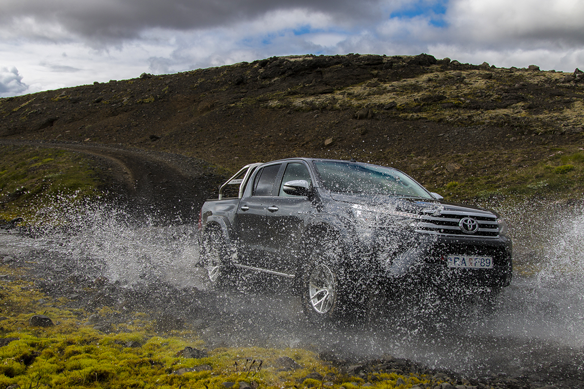 A black pickup truck drives through a shallow river with splashing water, set against a rugged hillside under a cloudy sky, conveying adventure.