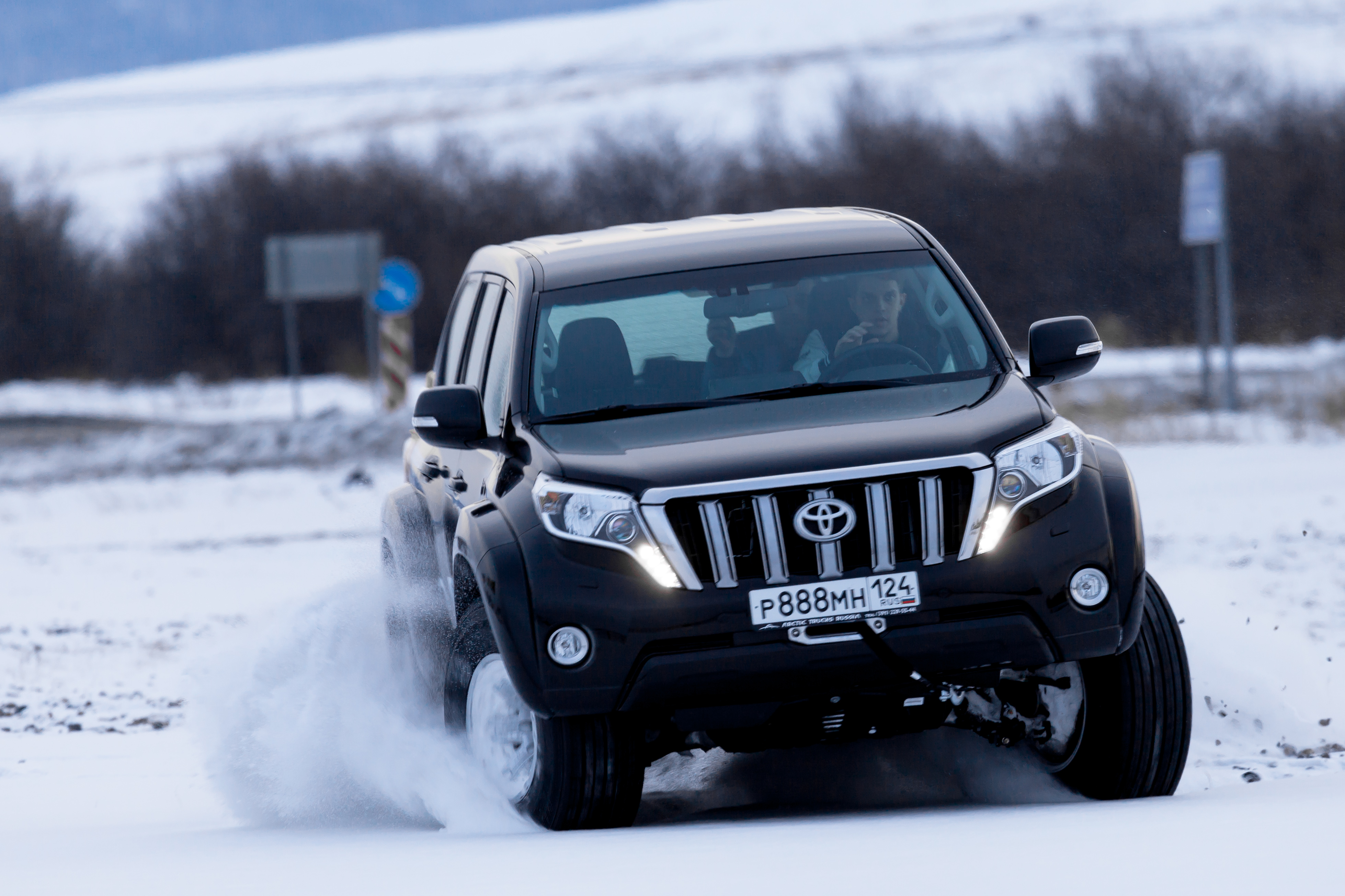 A black Toyota SUV skids on a snow-covered road, creating a dynamic spray of snow. The background features blurred trees and overcast skies, conveying a sense of speed and winter adventure.