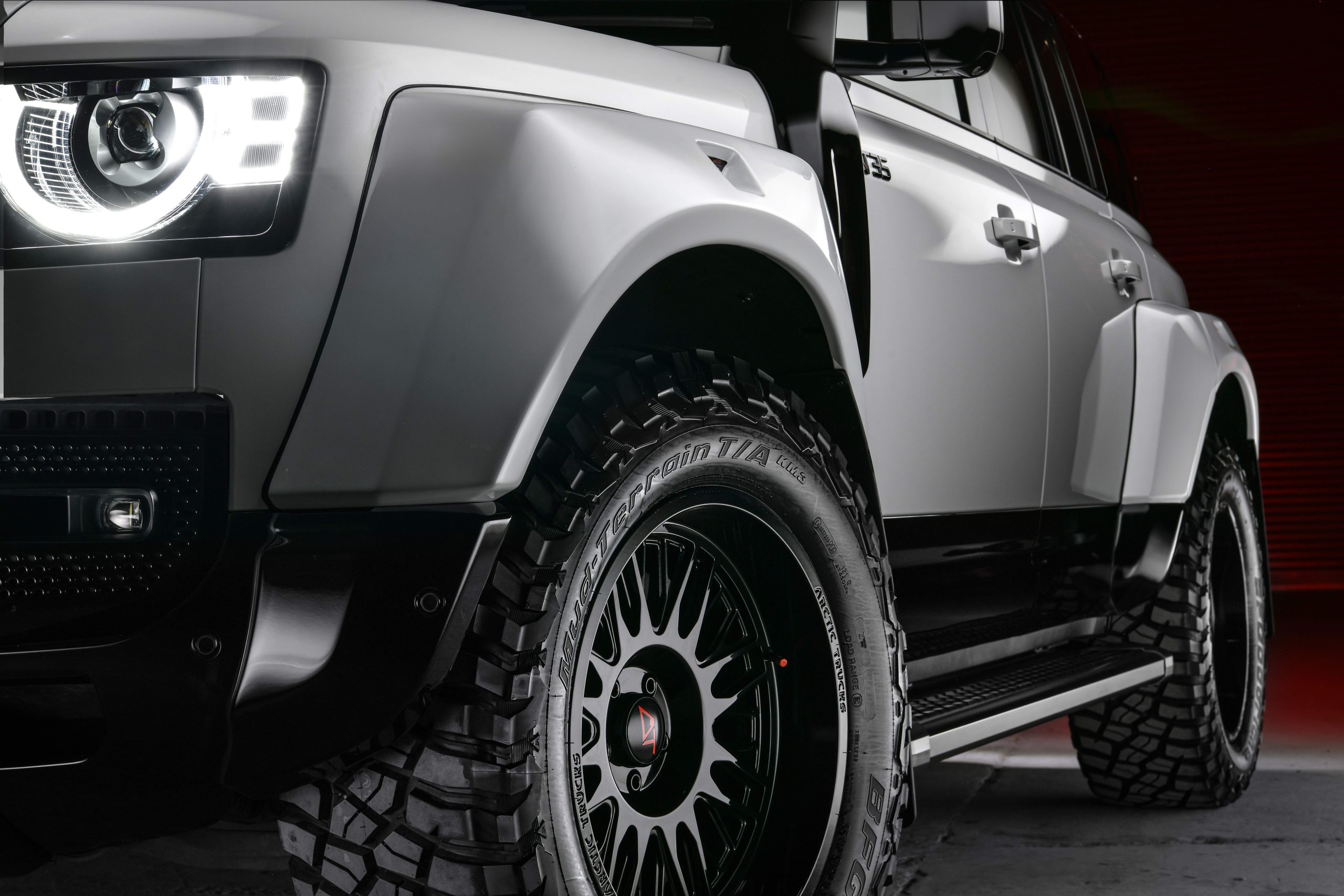 Close-up of a rugged SUV with illuminated circular headlight, matte silver body, and large black off-road tires, set against a dark red background.
