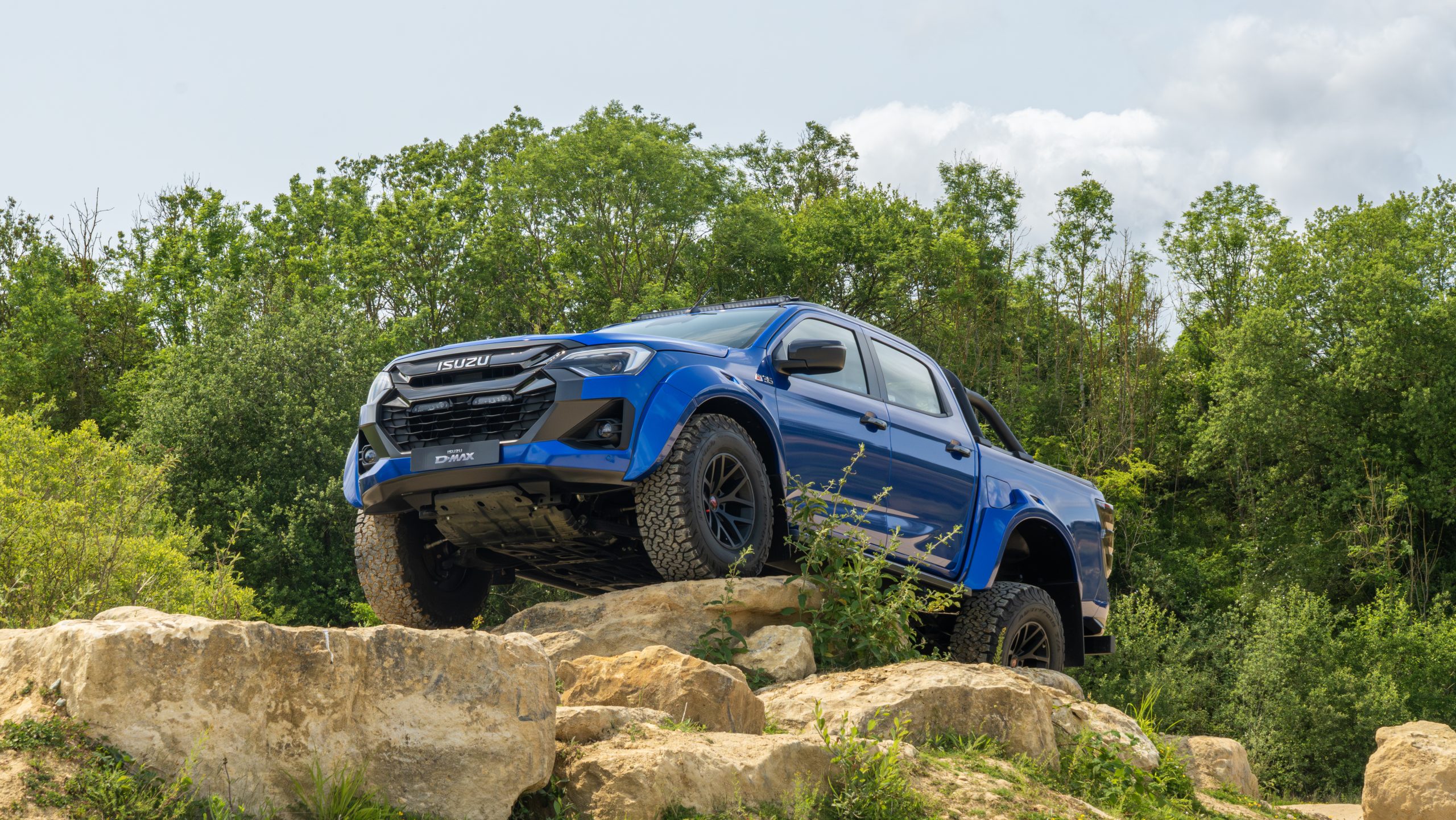 A blue Isuzu pickup truck is parked boldly on large rocks in a forested area. The scene conveys adventure and rugged capability against a backdrop of green trees and a partly cloudy sky.