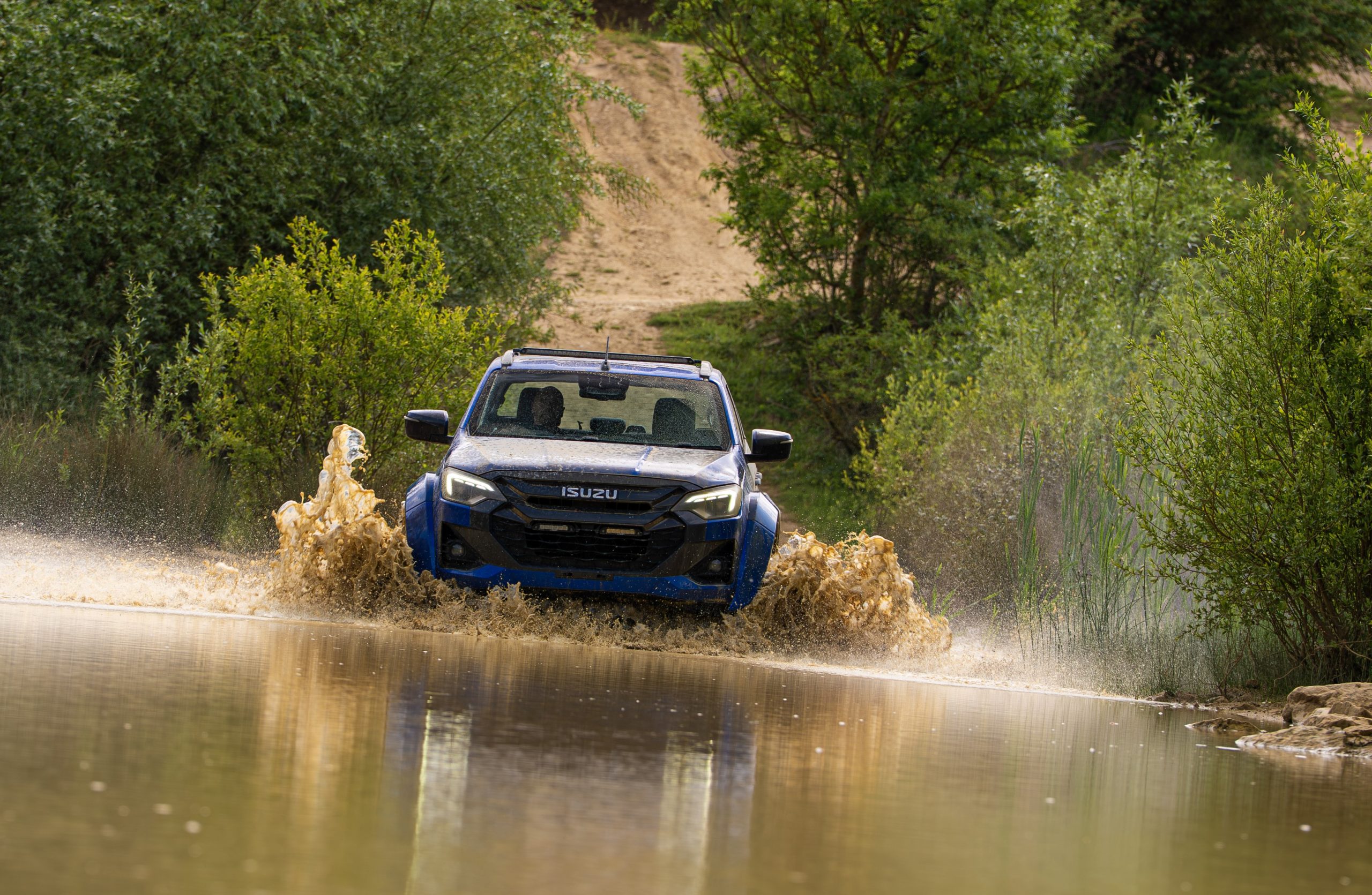 A blue Isuzu SUV splashes through a muddy water trail flanked by lush green bushes, creating a dynamic and adventurous scene.