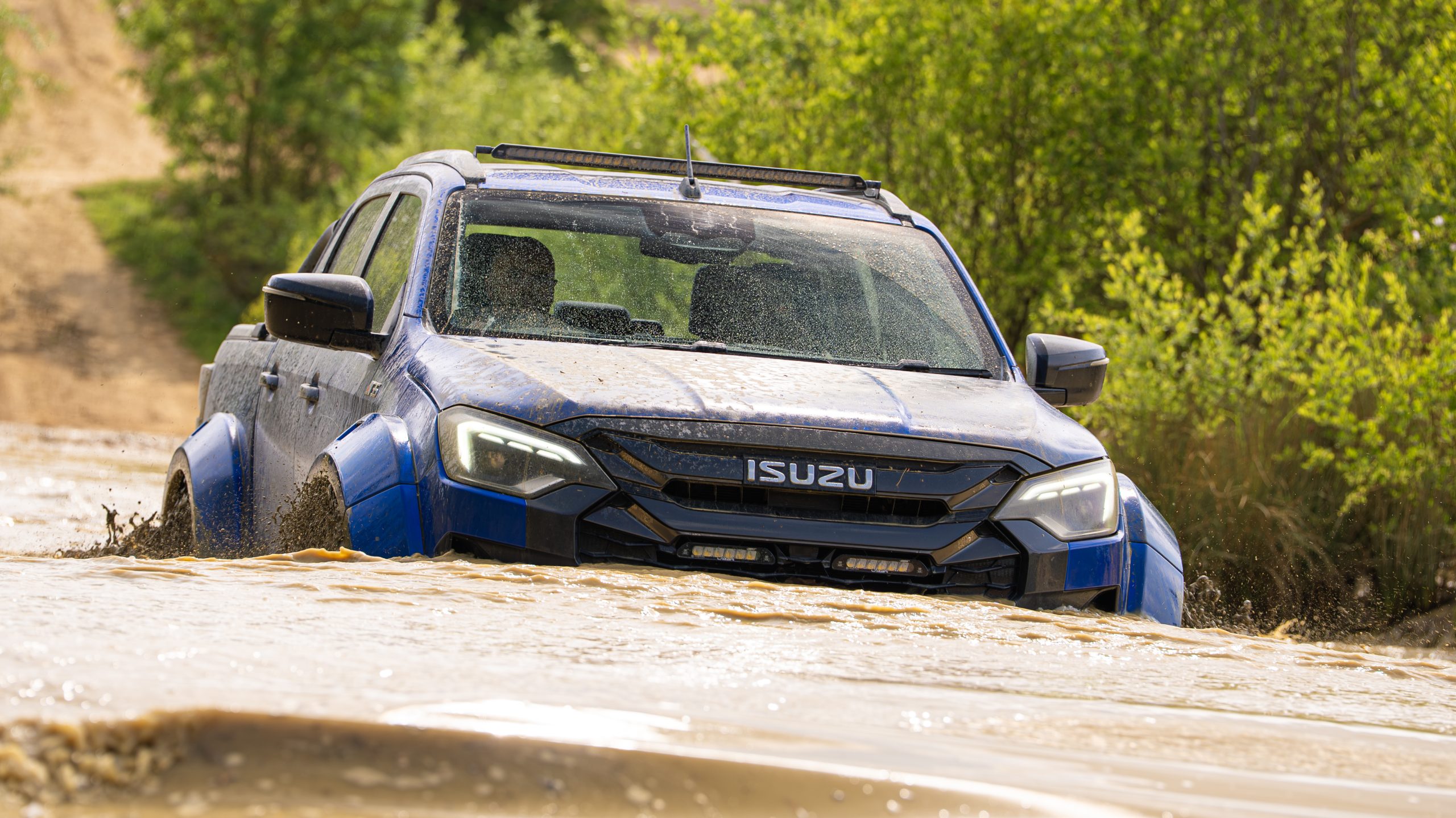 A blue Isuzu truck fords through muddy water, splashing around its rugged body. The background shows lush green foliage and a dirt track.