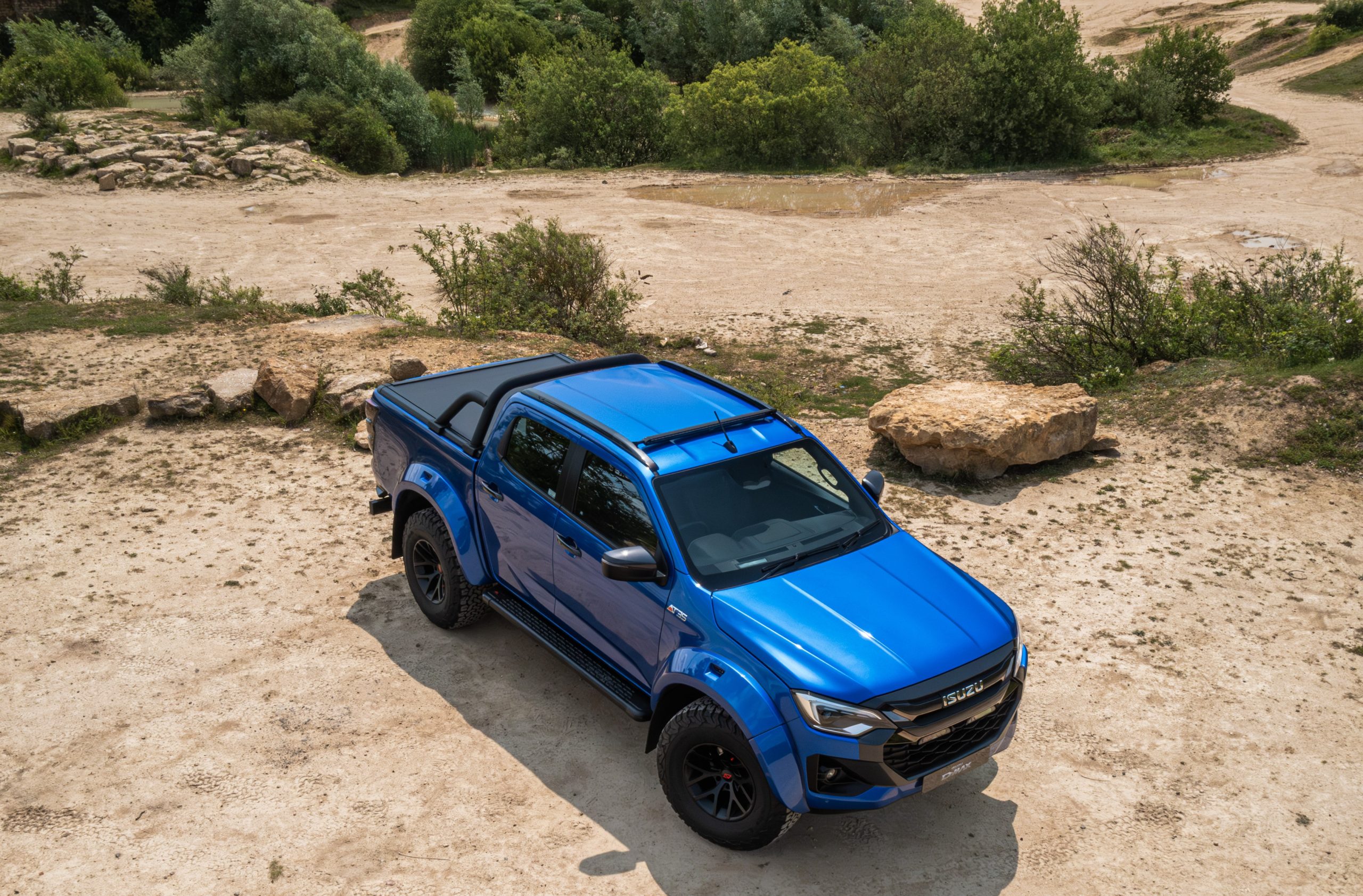 A vibrant blue pickup truck is positioned on a rugged, sandy terrain with scattered rocks and lush green bushes in the background, conveying adventure.