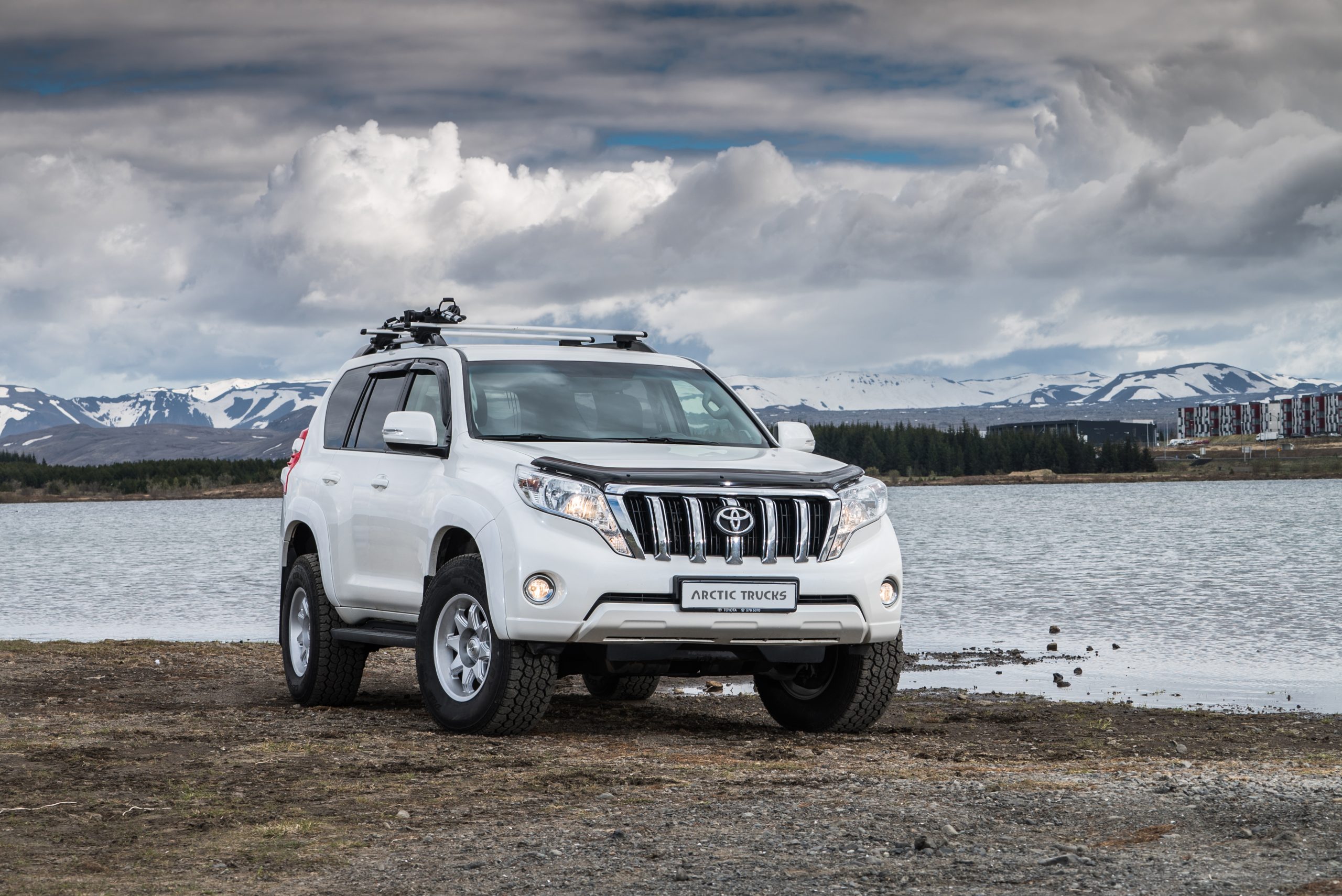 White SUV parked by a serene lake, with snow-capped mountains and a cloudy sky in the background, conveying an adventurous and rugged tone.