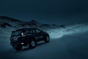 A rugged SUV navigates a snow-covered road under a dark, moody sky in a barren, mountainous landscape, conveying a sense of adventure and solitude.