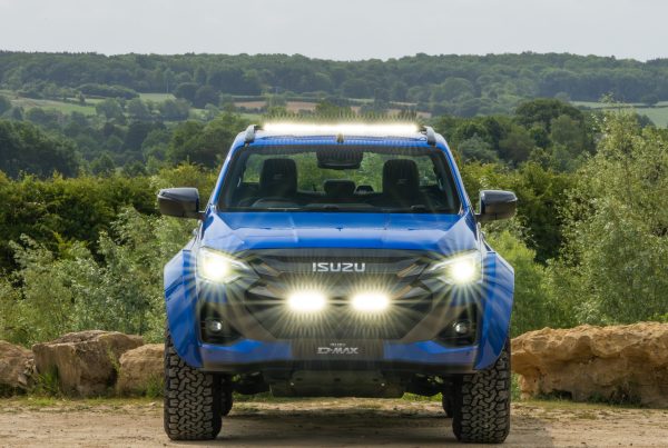Blue Isuzu D-Max pickup with bright headlights on, parked on a dirt path. Dense green forest and rolling hills form the background under a cloudy sky.