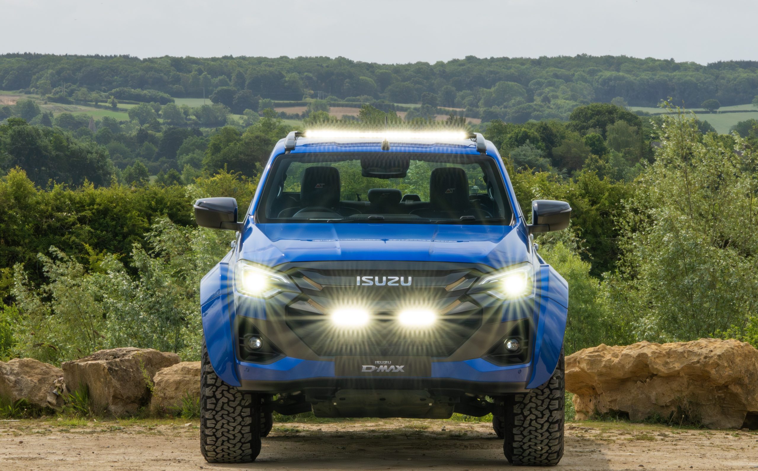 Blue Isuzu D-Max pickup with bright headlights on, parked on a dirt path. Dense green forest and rolling hills form the background under a cloudy sky.