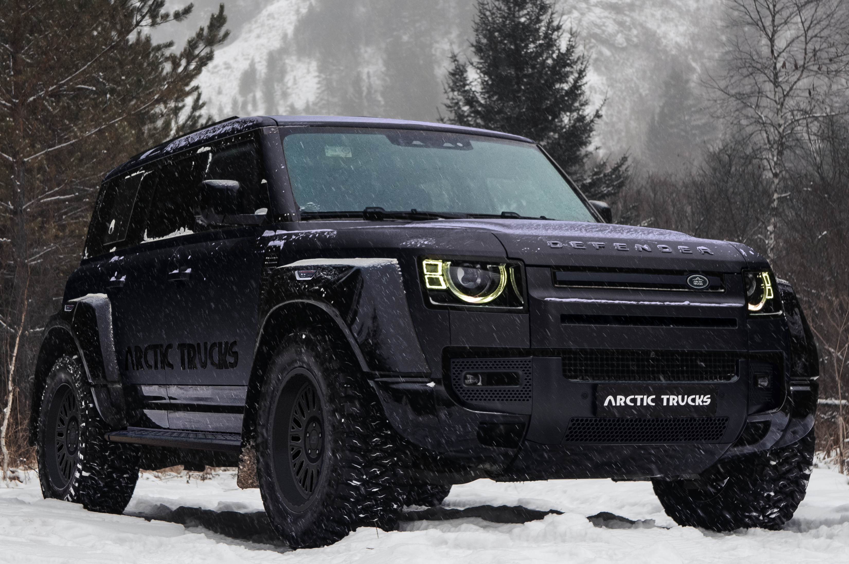 A black Arctic Trucks SUV with large tires is parked on snowy ground in a forest. Snow gently falls, conveying a rugged, adventurous tone.